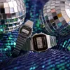 Two Casio digital watches are displayed on a sparkly blue and green sequined surface, with two disco balls in the background. Produktbild-Vorschau