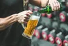 A person pouring beer into a glass mug in front of a rack of dumbbells. Produktbild-Vorschau