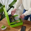 A person is putting money into a green cash box on a wooden table. Náhled obrázku produktu