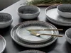A set of stoneware plates and bowls with a speckled gray and white glaze and a brown rim, arranged on a dark surface. Náhľad obrázku produktu