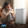 A woman sits on the floor next to a white oil radiator, looking down. Produktbild-Vorschau
