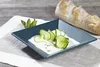 A square blue plate with cucumber slices and a creamy dip, garnished with mint leaves, sits on a white striped placemat next to a baguette on a wooden table. Produktbild-Vorschau