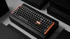 A black mechanical keyboard with orange accents is displayed on a tiered black platform against a gray background. Voir les images du produit