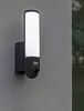 A modern outdoor security light with a built-in camera mounted on a textured gray wall. Produktbild-Vorschau