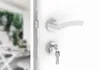 A close-up view of a white door with a silver door handle and lock mechanism, with keys inserted into the lock. The background is blurred and shows a dining area. Produktbild-Vorschau