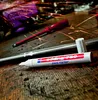 A paint marker and a screwdriver on a wooden surface with sparks flying in the background. Produktbild-Vorschau