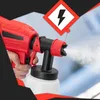 A hand holding a red and black electric paint sprayer, spraying a fine mist. A warning symbol for electricity is visible in the upper right corner. Produktbild-Vorschau
