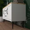 A white cabinet with black handles and legs, placed on a wooden floor in front of a green floral wallpaper. Produktbild-Vorschau