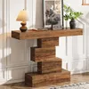 A narrow console table with a stacked block design is placed against a wall with white paneling. A lamp, a framed picture, and a vase with greenery are on top of the table. Anteprima dell’immagine prodotto