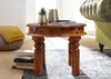 A round wooden coffee table with decorative legs, placed on a rug next to a green sofa and a basket with a yellow blanket. Produktbild-Vorschau