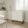A modern kitchen scene with white cabinets, a sink, a window with sheer curtains, and a white cabinet with black legs. A white dining table and chair are partially visible in the foreground. Voir les images du produit