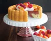 A cake on a cake stand with fruit and cream on top, with a basket of fruit in the foreground. Produktbild-Vorschau