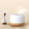 A white aroma diffuser with a wood-grain base is shown on a light-colored surface, with a small white vase containing a single white flower to its left. A plume of mist rises from the top of the diffuser. Anteprima dell’immagine prodotto