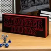 A Stranger Things logo light is displayed on a wooden surface, with dice and a miniature figure in the foreground. Produktbild-Vorschau