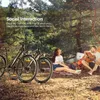 A couple is enjoying a picnic in a forest setting with two electric bicycles parked nearby. The man is playing a guitar while the woman looks up at the trees. Produktbild-Vorschau