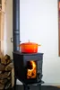 A red and orange enameled cast iron pot sits on top of a black wood-burning stove with a fire burning inside. Produktbild-Vorschau