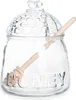 A clear glass honey jar with a honeycomb lid and a wooden honey dipper inside. Náhľad obrázku produktu