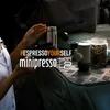 A person's hands are shown near a Wacaco Minipresso GR2 portable espresso machine, coffee beans in a jar, and a thermos, suggesting an outdoor or travel setting. Produktbild-Vorschau