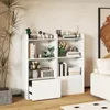 A white bookshelf with various decorative items and books on display, set in a room with a plant and a painting in the background. Produktbild-Vorschau