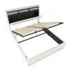 A white upholstered bed frame with the mattress support mechanism lifted to reveal storage space underneath. The headboard has a black panel with some compartments. Podgląd zdjęcia produktu