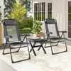 Two gray folding chairs and a small black table are set up on a patio with a rug underneath. There are plants and trees in the background, and a white building with windows behind the chairs. Produktbild-Vorschau