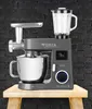 A multi-functional kitchen machine with various attachments is displayed against a dark brick wall background, placed on a wooden surface. Produktbild-Vorschau