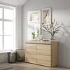 A wooden dresser with multiple drawers is placed against a light gray wall next to a large window. Two black wire baskets filled with blankets are on the floor in front of the dresser. Artwork and vases with branches are displayed on top of the dresser. Th Produktbild-Vorschau