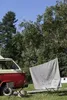 A red and beige camper van is parked in a grassy area with trees in the background. A grey blanket with a mountain graphic and text is hung on a clothesline between two chairs. A small pillow with a similar graphic is on one of the chairs. Produktbild-Vorschau