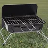 A black portable grill with silver legs and handles sits on a grassy lawn. Produktbild-Vorschau