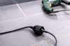 A black extension cord with a connector is plugged into another black cord, which is connected to a green power drill. The items are on a tiled floor. Produktbild-Vorschau