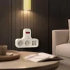 A white power strip with a red switch is mounted on a dark wooden wall, illuminated by a yellow wall lamp. A sofa and a coffee table with a book and a mug are visible in the foreground. Produktbild-Vorschau