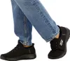 A person wearing blue jeans and black Skechers slip-on shoes. Voir les images du produit