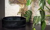 A black basin sink with a gold faucet is shown on a dark countertop, with a large green plant and a textured wall in the background. Produktbild-Vorschau