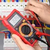A person is using a red digital multimeter to test electrical components in a breaker box. Podgląd zdjęcia produktu