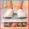 A person's feet wearing slippers and orange socks are standing on a storage container filled with building blocks. Náhľad obrázku produktu