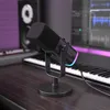 A black microphone is placed on a wooden desk in front of a keyboard, with a studio monitor speaker and a computer screen displaying audio waveforms in the background. The scene is lit with purple and blue ambient lighting. Produktbild-Vorschau