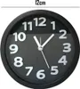 A close-up of a black analog alarm clock with white numbers and hands, set against a black background. A measurement of 12cm is indicated above the clock. Produktbild-Vorschau