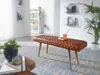 A brown leather bench with diamond stitching is placed in a living room setting, with a plant and a coffee table in the foreground. Produktbild-Vorschau