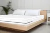 A white mattress topper with a black stripe is placed on a wooden bed frame, with two white pillows at the head of the bed and a potted plant to the left. Produktbild-Vorschau