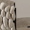 A close-up view of a textured beige fabric product, possibly a stool or chair, with a black frame visible. Another piece of the same fabric is in the foreground with a small black label. Produktbild-Vorschau