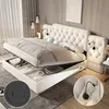 A bedroom scene featuring a large, tufted beige bed with its storage compartment open, revealing stored items. Two inset circular images highlight a USB/power outlet and a reading lamp. Produktbild-Vorschau