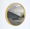 A round gold-colored mirror is mounted on a white wall, reflecting a staircase and a plant. Náhled obrázku produktu