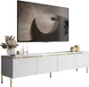 A white TV cabinet with gold accents and legs, featuring a large flat-screen TV mounted above it. Decorative items like a vase with dried flowers, candles, and spheres are placed on top of the cabinet. Náhled obrázku produktu
