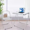 A transparent acrylic folding chair and a white folding desk with a laptop are shown in a bright room with blinds. Produktbild-Vorschau