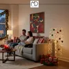 A couple sits on a couch in a living room decorated for Christmas, looking at a tablet. A decorative light tree with ornaments stands nearby, with gift boxes at its base. Podgląd zdjęcia produktu