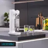 A white Arendo soda maker with a bottle filled with carbonated water is placed on a dark countertop next to two glasses with lime slices. In the background, a modern kitchen setting is visible with a faucet, cutting boards, and a bowl of lemons. Produktbild-Vorschau