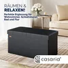 A black storage bench in a living room setting, with text overlays and a brand logo. Produktbild-Vorschau