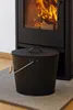 A black metal ash bucket with a lid and handle sits on a protective mat in front of a black wood-burning stove with a fire burning inside. Produktbild-Vorschau