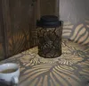 A black metal lantern with a leaf pattern is placed on a textured surface next to a white cup of coffee. The lantern casts intricate leaf-shaped shadows on the surface and a wooden wall behind it. Produktbild-Vorschau