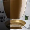 A close-up shot of a gold-colored plastic planter with a matching saucer, set against a bright, modern interior background with a snake plant in a white planter visible. Produktbild-Vorschau